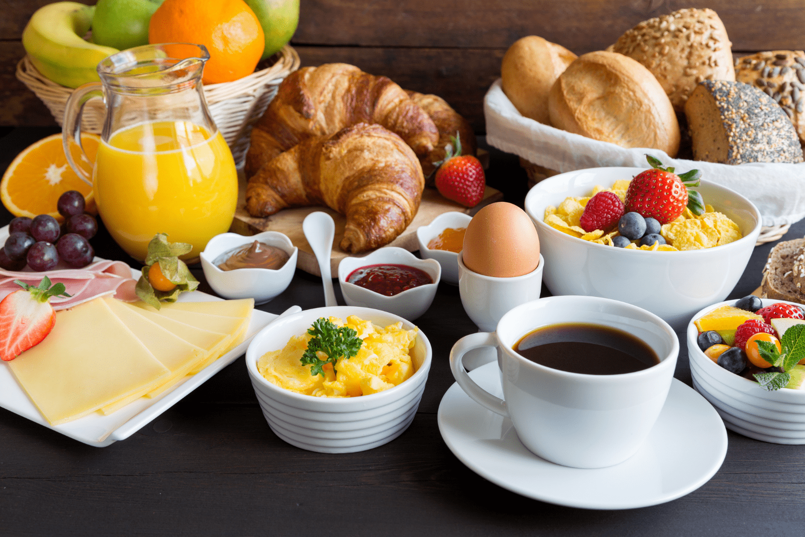 A breakfast spread on a table with scrambled eggs, a boiled egg, black coffee, orange juice, croissants, bread rolls, sliced cheese and ham, bowls of cereal topped with berries, fresh fruit salad, grapes, strawberries, and small dishes of jam and spreads.
