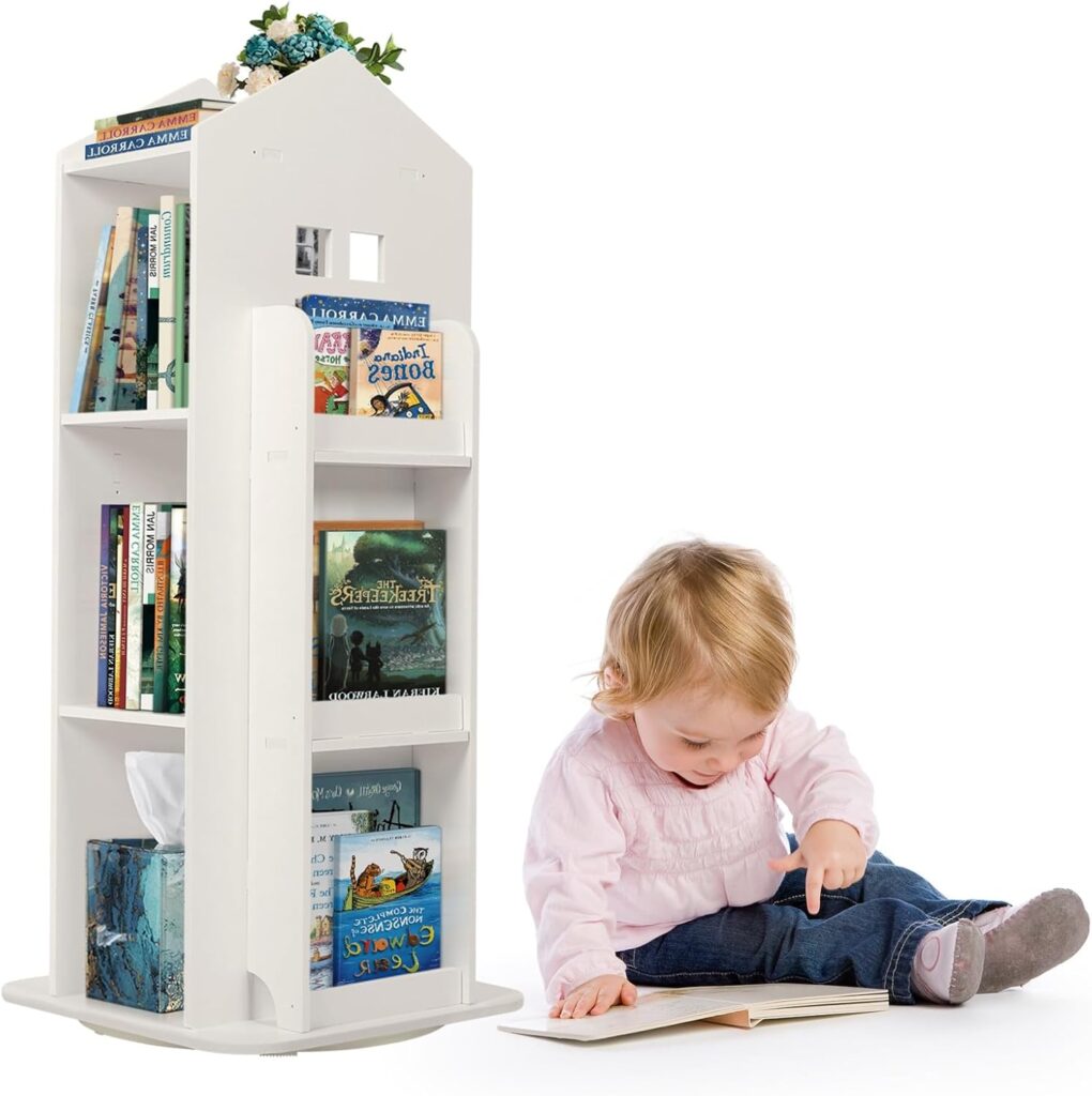 A young child sits on the floor reading a book next to a white, house-shaped rotating bookshelf filled with children’s books. The bookshelf has multiple shelves, some displaying book covers, and a tissue box on the bottom shelf. A small bouquet of flowers decorates the top. The background is white and brightly lit.